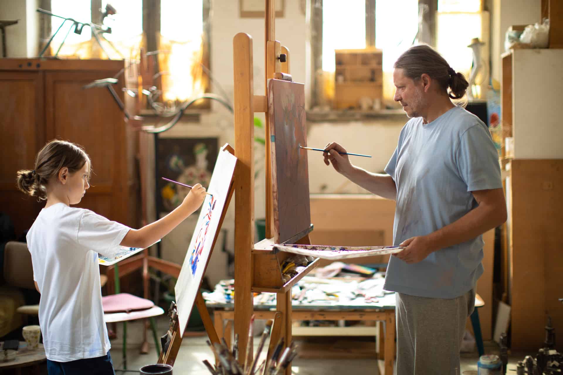 Man and boy painting together in artist studio. Both of them has they own canvas. They wear casual clothes and both of them have long hair. Studio is full with artworks, paints and sculptures. Property released.